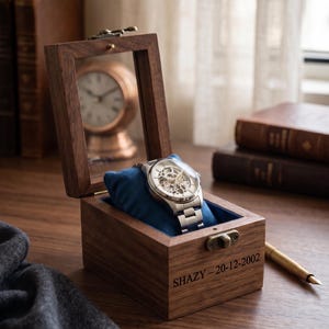 May include: A silver wristwatch with a skeleton dial rests on a blue velvet cushion inside a wooden watch box. The box is engraved with the name "SHAZY" and the date "20-12-2002". The box is open, revealing the watch, and is set on a wooden surface.