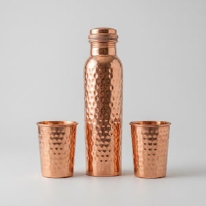 Puede incluir: Una botella de agua de cobre y dos vasos a juego, todos con textura martillada. La botella tiene una tapa de rosca. El juego se muestra sobre un fondo blanco.