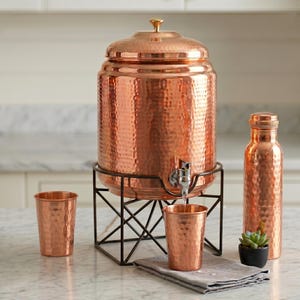 May include: A copper water dispenser with a hammered texture, a matching water bottle, and two copper cups. The dispenser sits on a black metal stand. A small succulent in a black pot is next to the bottle.