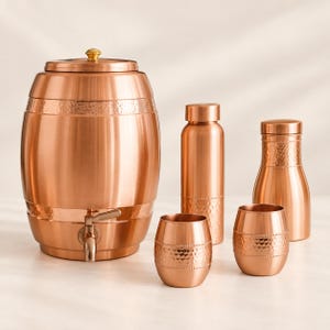 May include: A copper beverage dispenser with a spigot and lid, accompanied by a tall bottle, a smaller carafe, and two matching tumblers. Each item features a polished metallic finish with a decorative hammered texture band around the center.