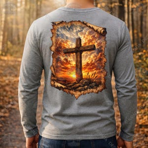 May include: A gray long-sleeve shirt featuring a detailed graphic of a wooden cross against a sunset backdrop. The artwork is framed within a torn paper effect, creating a unique visual appeal. The shirt is a comfortable, everyday wear item.