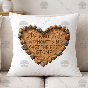 May include: White square pillow with a heart-shaped design. The heart is made of brown stone and outlined with small rocks. The text "HE WHO IS WITHOUT SIN CAST THE FIRST STONE" is carved into the stone heart.