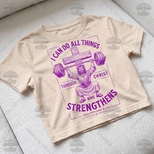 May include: A beige t-shirt featuring a purple graphic of Jesus lifting a barbell made of a cross. The text reads "I can do all things through Christ who strengthens" and "Philippians 4:13".