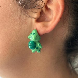 May include: A small, glossy, light green earring shaped like a stylised creature with a dark green bulb-like feature on its back and darker green spots on its body. The earring has a smooth, polished finish and is worn on the earlobe.