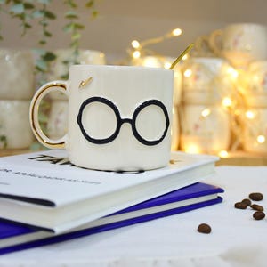 May include: A white ceramic mug with black glasses design and a gold handle, resting on a stack of books. A gold spoon is inside the mug. Coffee beans are scattered on the white surface. Other mugs are in the background.