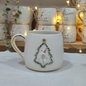 Puede incluir: Taza de cerámica blanca con asa dorada. La taza presenta un diseño de árbol de Navidad con un muñeco de nieve con sombrero y corona. Otras tazas similares están apiladas en el fondo, sugiriendo un tema navideño.