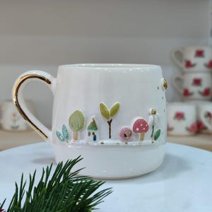 Peut inclure: Tasse en céramique blanche avec une anse et un bord dorés. La tasse est décorée d'une scène de jardin fantaisiste avec des arbres, des maisons et des champignons. La tasse repose sur un présentoir à gâteau en marbre blanc.
