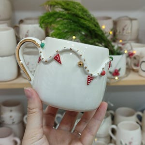 May include: A white ceramic mug with a gold handle and rim, decorated with a garland of red and green accents. The mug is held in a hand, with other mugs visible in the background, suggesting a holiday theme.