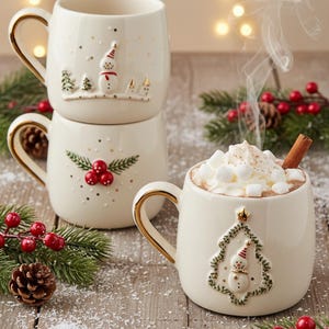 Op de afbeelding: Crèmekleurige keramische mokken met gouden handvatten en feestelijke ontwerpen. Eén mok is gevuld met warme chocolademelk, gegarneerd met marshmallows en een kaneelstokje. De andere mokken hebben sneeuwpop- en bessenmotieven, perfect voor feestelijke drankjes.