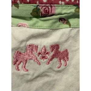 Pode incluir: Close-up de um detalhe de tecido com um desenho bordado em rosa. O desenho apresenta dois cavalos estilizados ladeando uma flor-de-lis. O tecido tem uma base branca com padrões florais e de bolinhas em rosa e verde.