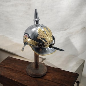 May include: A silver-colored metal helmet with a prominent central spike and a gold-colored eagle emblem. The helmet features a decorative gold band and is displayed on a wooden stand, showcasing historical military headwear.