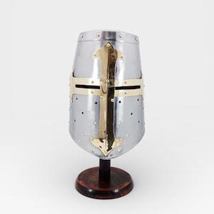 May include: A silver medieval helmet featuring a gold cross design. The helmet is displayed on a dark brown wooden stand. The helmet has small ventilation holes, and is a historical item. The stand is made of wood.