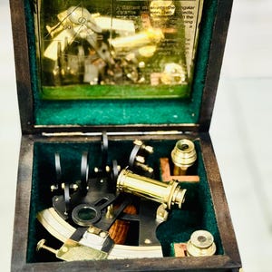 Könnte beinhalten: Ein Vintage-Sextant in einer Holzkiste. Die Kiste ist dunkelbraun mit Messingbeschlägen und einem grünen Samtinterieur. Der Sextant ist aus Messing gefertigt und hat eine Lupe und andere Komponenten. Der Text "WHAT DOES A SEXTANT DO?" ist sichtbar.
