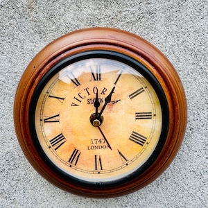 Puede incluir: Un reloj redondo de madera con marco marrón claro y anillo interior negro. La esfera es de color crema vintage con números romanos negros y las palabras "VICTOR STATION 1747 LONDON". Las manecillas son negras.