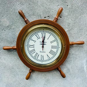 Könnte beinhalten: Eine Holzschiff-Rad-Uhr mit weißem Zifferblatt und römischen Ziffern. Die Uhr hat eine messingfarbene Lünette und Holzspeichen. Das Zifferblatt zeigt "London" und den Markennamen. Die Uhr ist an einer grauen Wand befestigt.