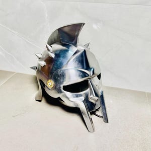 Könnte beinhalten: Ein silberfarbener Gladiatorenhelm mit einem markanten Kamm und spitzen Details. Der Helm hat einen Gesichtsschutz und ein dekoratives goldfarbenes Emblem. Der Metallhelm wird auf einer hellen Oberfläche präsentiert, die sein Design hervorhebt.