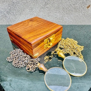 Può includere: Una scatola quadrata in legno con chiusura e cerniere in ottone, accompagnata da due lenti d'ingrandimento su lunghe catene in metallo dorato. La scatola presenta una venatura del legno visibile e le lenti hanno lenti rotonde e manici in metallo.