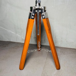 May include: A wooden tripod with three legs, featuring a silver metal top and accents. The legs are a warm brown colour, and a silver chain connects the legs near the bottom. The tripod is designed for supporting equipment.
