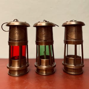 Op de afbeelding: Drie antieke messing lantaarns met rode, groene en heldere glazen panelen. Elke lantaarn heeft een cirkelvormig embleem met de tekst "Flame for Royal Navy London 1920". De lantaarns hebben een handvat en een voet.