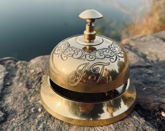 May include: A polished brass bell with a decorative floral pattern and the initials "J.T." engraved on the surface. The bell has a rounded top with a small handle and sits on a circular base. The background is blurred, with a body of water visible.
