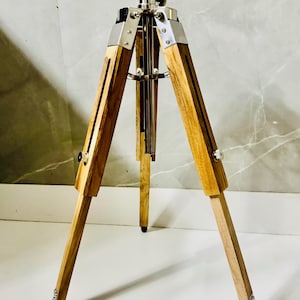May include: A wooden tripod with a silver metal top and accents. The tripod has three wooden legs with metal details and black feet. The legs are adjustable. The tripod is designed for photography or display.