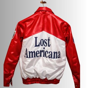 May include: A red and white satin jacket with the words "Lost Americana" in navy blue lettering on the back. The jacket has a classic bomber style with a ribbed hem and cuffs. Patches are visible on the sleeves.