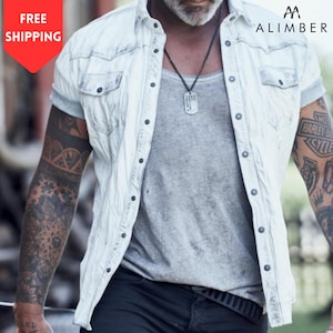 May include: A light-wash denim shirt with short sleeves, a button-down front, and two chest pockets. The shirt is worn over a grey vest. The image also shows a black necklace and tattoos on the arms. The brand name "ALIMBER" is visible.