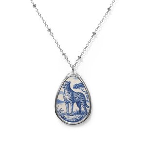 Puede incluir: Collar plateado con un colgante en forma de lágrima. El colgante muestra una ilustración azul y blanca de un guepardo en un paisaje con un árbol. El colgante tiene un marco plateado y una cadena delicada.