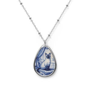Puede incluir: Collar plateado con un colgante en forma de lágrima que presenta una ilustración de un gato siamés. El gato está representado en un diseño azul y blanco, que recuerda a la cerámica de Delft. El colgante cuelga de una delicada cadena.