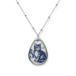 May include: Silver-toned necklace with a teardrop pendant featuring a blue and white illustration of a seated cat. The cat has blue stripes and is set against a background with a house and trees.