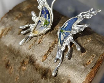 May include: A pair of silver-toned butterfly earrings. Each earring features a blue and white floral design encased in a silver frame. The wings have a unique, flowing shape, and the earrings are displayed on a piece of wood.