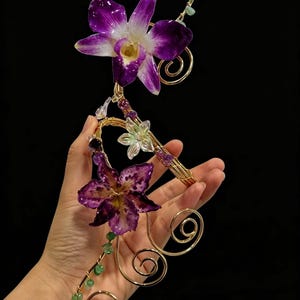 Peut inclure: Une boucle d'oreille en fil doré ornée de deux grandes fleurs d'orchidée violettes, d'une petite fleur translucide et de petites perles en pierre verte et violette. Le fil métallique est formé en spirales et courbes décoratives.