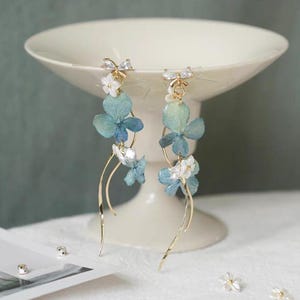 Könnte beinhalten: Elegante Hängeohrringe mit blauen und weißen floralen Akzenten. Die Ohrringe haben goldfarbene Reifen und zarte, fließende Linien. Klare Kristalle verleihen dem Design Glanz. Präsentiert auf einem weißen Podest.
