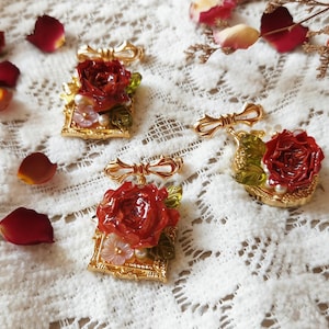 Puede incluir: Cuatro broches dorados con detalles de rosas rojas y perlas. Cada broche presenta un detalle de lazo en la parte superior y se coloca sobre un fondo de encaje blanco con pétalos de rosa esparcidos. Los broches miden aproximadamente 3,8 cm de largo.