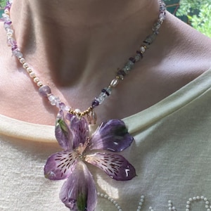 Op de afbeelding: Een ketting met een grote hanger van een geperste paarse alstroemeria-bloem. De ketting bestaat uit kleine witte parels, doorzichtige kralen en onregelmatige paarse amethistkleurige steentjes, geaccentueerd met kleine goudkleurige kraaltjes.