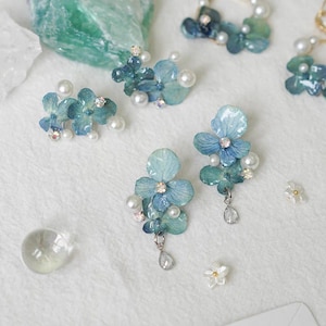 Könnte beinhalten: Nahaufnahme von mehreren Schmuckstücken, darunter Ohrringe und ein Anhänger, mit blau-grünen Blumendesigns mit Perlenakzenten und kleinen Kristallen. Die Ohrringe haben einen Blumen- und Perlencluster.