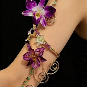 May include: An arm cuff featuring gold-toned wire spirals, two large purple orchid-like flowers, a smaller translucent white flower, and small green and purple gemstone accents. The wire structure wraps around the arm with floral embellishments.