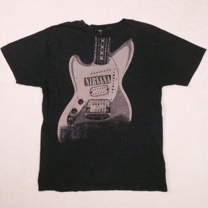 Puede incluir: Camiseta negra con un gráfico gris de una guitarra y el nombre de la banda "NIRVANA" impreso. El diseño de la guitarra es una ilustración estilizada. La camiseta está hecha de un material suave.