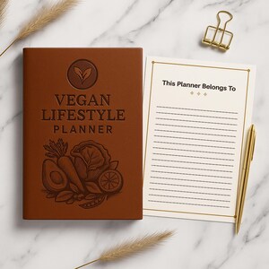 Peut inclure: Un agenda marron "Vegan Lifestyle Planner" avec du texte en relief et des illustrations de légumes. L'agenda comprend une page lignée avec le texte "This Planner Belongs To" et un stylo doré. Un trombone doré et une plante décorative sont également présents.