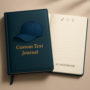 Peut inclure: Un journal bleu canard avec un motif de casquette de baseball et l'inscription dorée "Custom Text Journal". Le journal ouvert révèle des pages lignées et le texte "A5 NOTEBOOK". Un signet est visible.