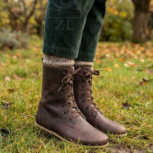 May include: Brown leather lace-up boots with a round toe and a flat sole. The boots are mid-calf height and have brown laces. The boots are worn with green corduroy pants and tan socks. The boots are on a grassy surface.