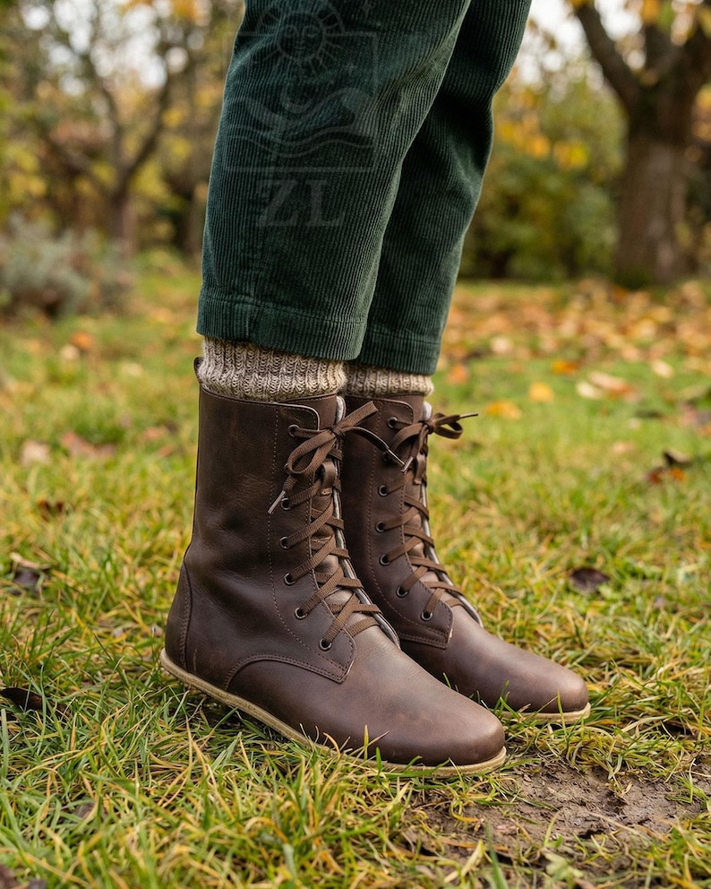 May include: Brown leather lace-up boots with a round toe and a flat sole. The boots are mid-calf height and have brown laces. The boots are worn with green corduroy pants and tan socks. The boots are on a grassy surface.