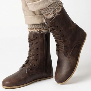 May include: Brown leather boots with a lace-up design and side zipper. The boots have a flat sole and are worn with beige pants and patterned socks. The boots are a dark brown color.