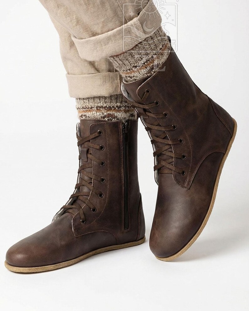 May include: Brown leather boots with a lace-up design and side zipper. The boots have a flat sole and are worn with beige pants and patterned socks. The boots are a dark brown color.