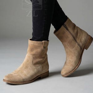 May include: Tan suede ankle boots with a perforated design on the upper shaft. The boots have a side zipper and a low stacked heel. The boots are worn with black jeans. The boots have a round toe.