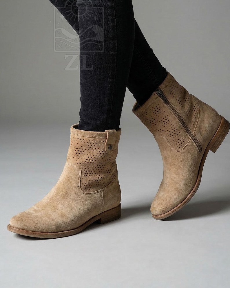 May include: Tan suede ankle boots with a perforated design on the upper shaft. The boots have a side zipper and a low stacked heel. The boots are worn with black jeans. The boots have a round toe.