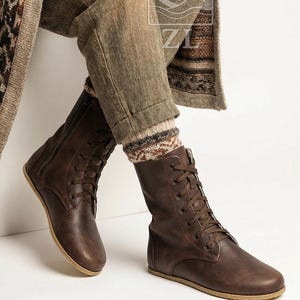 May include: Brown leather lace-up boots with a side zipper. The boots have a flat sole and are worn with patterned socks and brown pants. The boots are a medium brown color and appear to be made of a soft leather material.