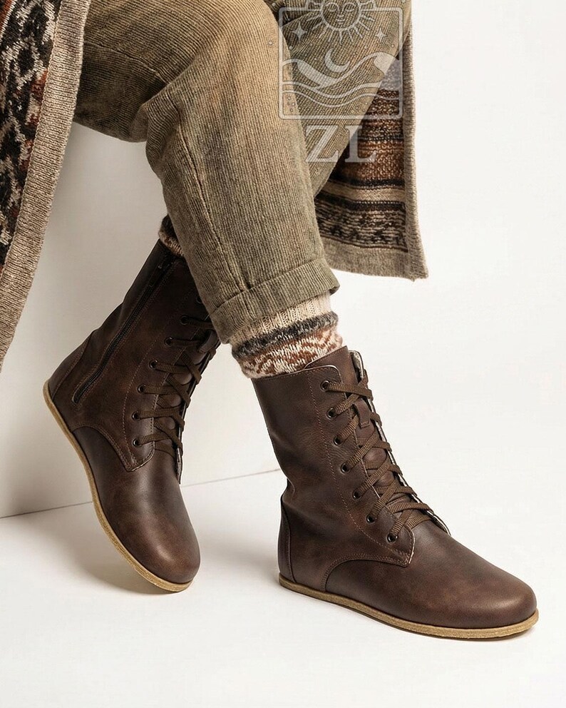 May include: Brown leather lace-up boots with a side zipper. The boots have a flat sole and are worn with patterned socks and brown pants. The boots are a medium brown color and appear to be made of a soft leather material.