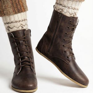 May include: Brown leather lace-up boots with side zippers. The boots have a flat sole and are worn with patterned socks and brown pants. The boots are a dark brown color and appear to be made of leather.