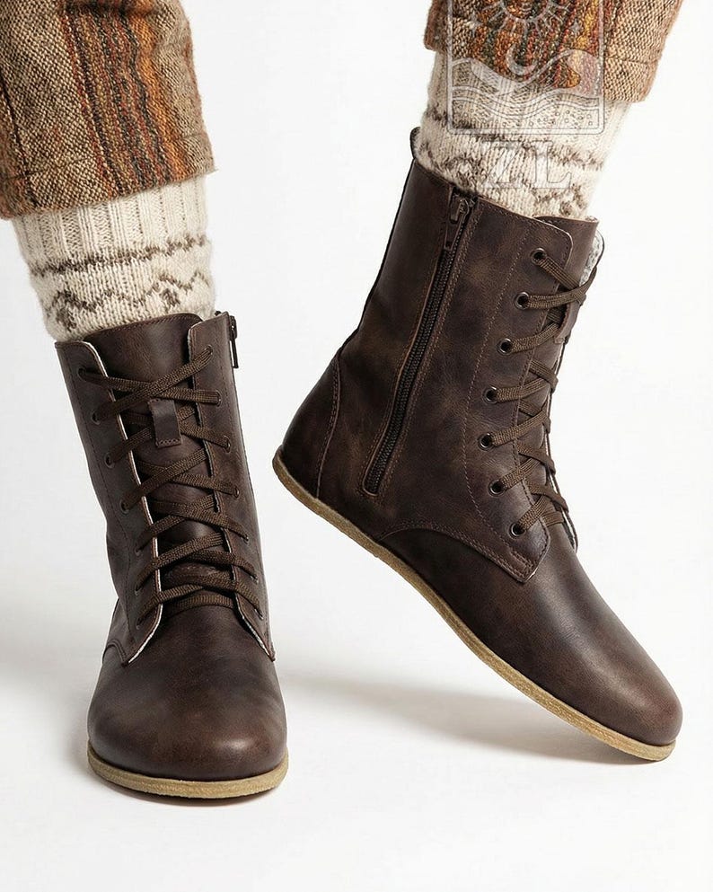 May include: Brown leather lace-up boots with side zippers. The boots have a flat sole and are worn with patterned socks and brown pants. The boots are a dark brown color and appear to be made of leather.